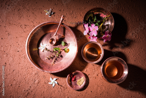 copper kalash, glass, spoon and plate used by Bramhins after sacred thread ceremony while performing Sandhya Vandanam or Sandhya Kriya, over clay background with tulsi or basil leaves & flowers

