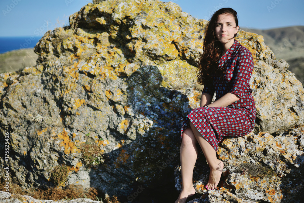 Naklejka premium Young beautiful girl sits on a stone, straight calm look. Emotions: waiting, willingness, patience