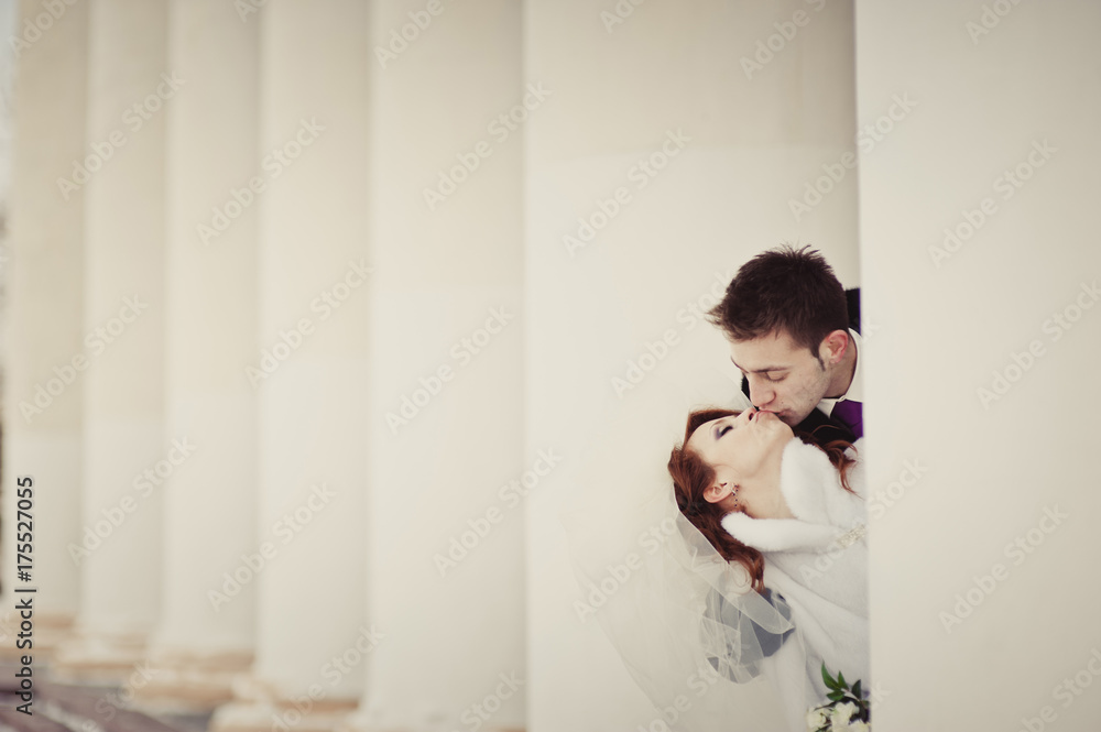 A beautiful ginger bride in a white fur coat and groom peeking out from behind the white columns. Winter wedding outdoors. A beautiful ginger bride in a white fur coat and groom peeking out from behind the white columns. Winter wedding outdoors.