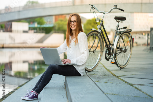 a young charming woman sitting near the river somewhere in the city and working with a laptop; her bicycle is standing next to her; 