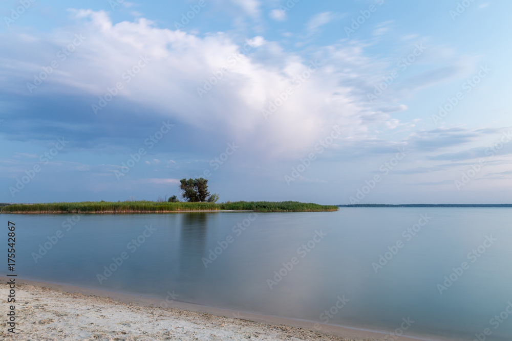 Beautiful scenic landscape of river or lake bank, calm water and island with tree against dramatic cloudy sky