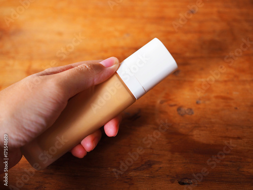 Left hand holding beige BB concealer foundation makeup cosmetic cream transparent bottle with white cap, with shadow, on red brown wooden texture table top background