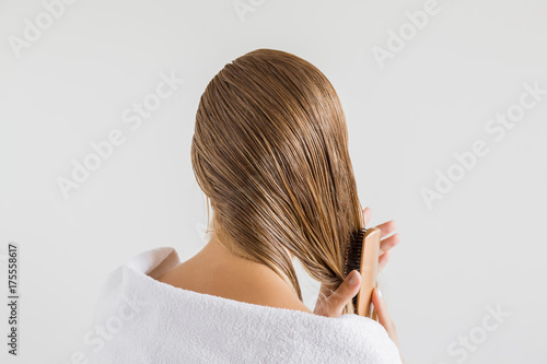 Woman in the white towel with comb brushing her wet blonde hair after shower on the gray background. Cares about a healthy and clean hair. Beauty salon concept.
