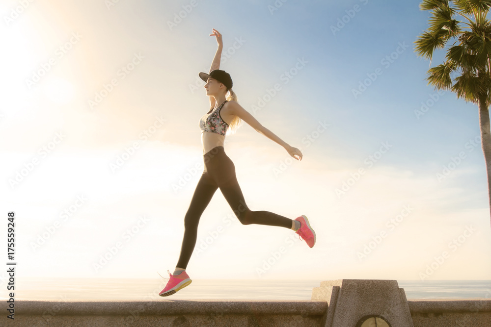 Fototapeta premium Beautiful young woman jumping and exercise close to the sea with sunset behind her. Golden colors made nice feeling and matching with long blonde hair of the girl. 