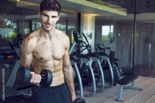 Young handsome muscular man exercising in the gym. Dumbbell bicep curl exercise in standing position next to the weight rack. Exercise with the smile.