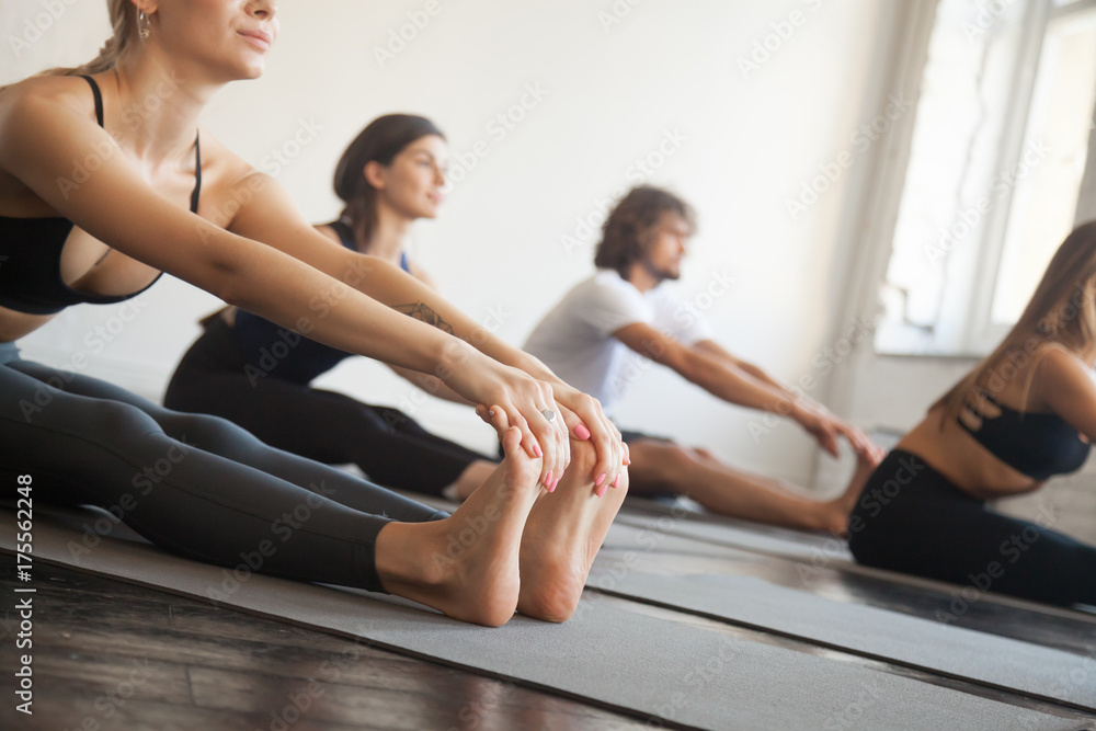 Group of young sporty people practicing yoga lesson with instructor concept, sitting in paschimottanasana exercise, Seated forward bend pose, friends working out in club, indoor full length, studio