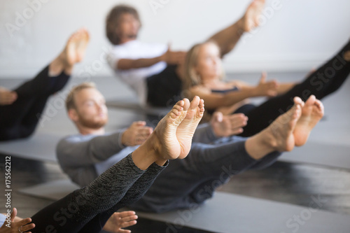 Group of young sporty people practicing yoga lesson with instructor, stretching in Paripurna Navasana exercise, balance pose, working out, indoor close up image, studio, focus on feet