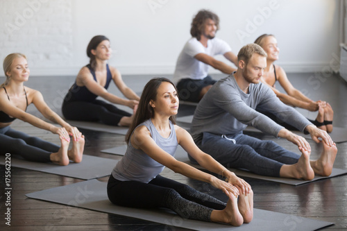 Group of young sporty people practicing yoga lesson with instructor, sitting in paschimottanasana exercise, Seated forward bend pose, indoor full length, studio, friends working out in club