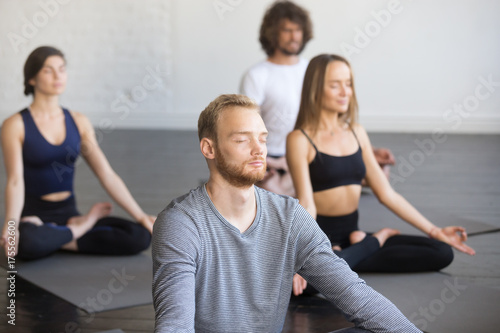 Group of young people, sporty students practicing yoga lesson with instructor, sitting and meditating with closed eyes in Padmasana exercise, Lotus pose, friends working out in club, studio