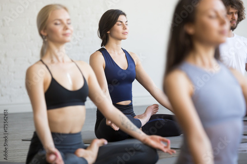 Group of sporty people, young students practicing yoga lesson with instructor, sitting and meditating with closed eyes in Padmasana exercise, Lotus pose, friends working out in club, studio background