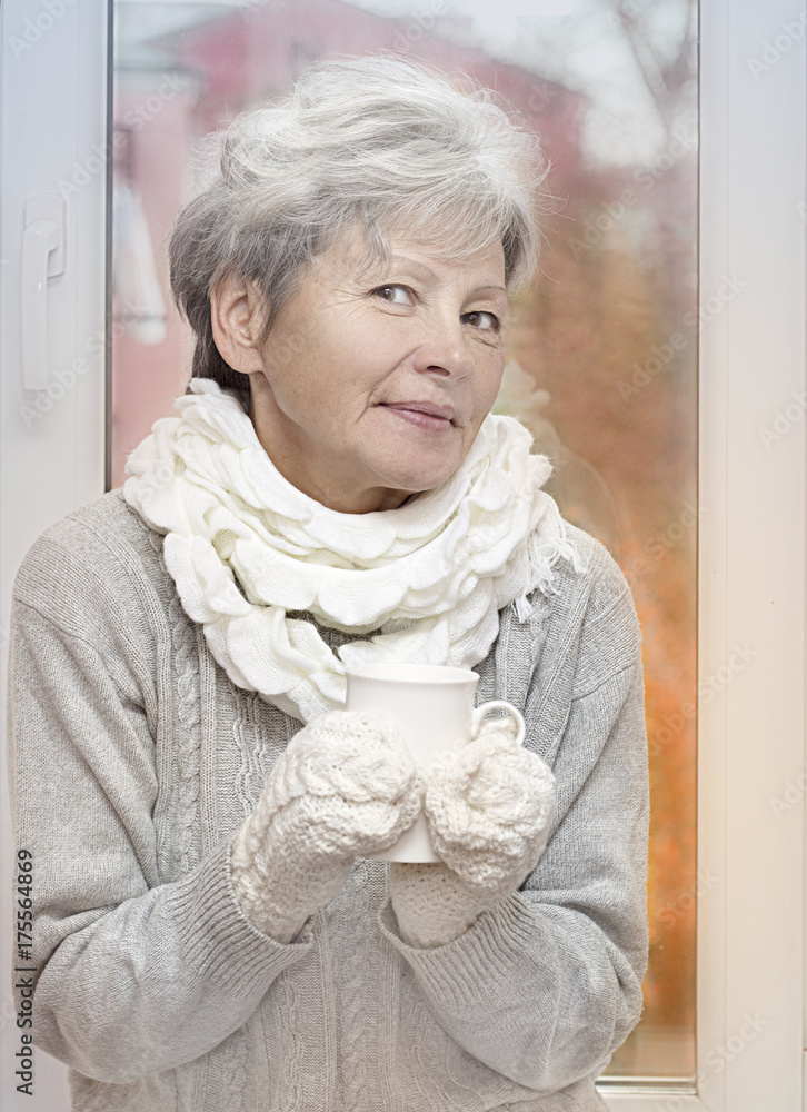 Gray haired woman in white knitted gauntlets drink beverage and smiling on autumn window background. Elderly woman with mug. Soft filter. Autumn toning effect. Gray haired woman in white knitted gauntlets drink beverage and smiling on autumn window background. Elderly woman with mug. Soft filter. Autumn toning effect.