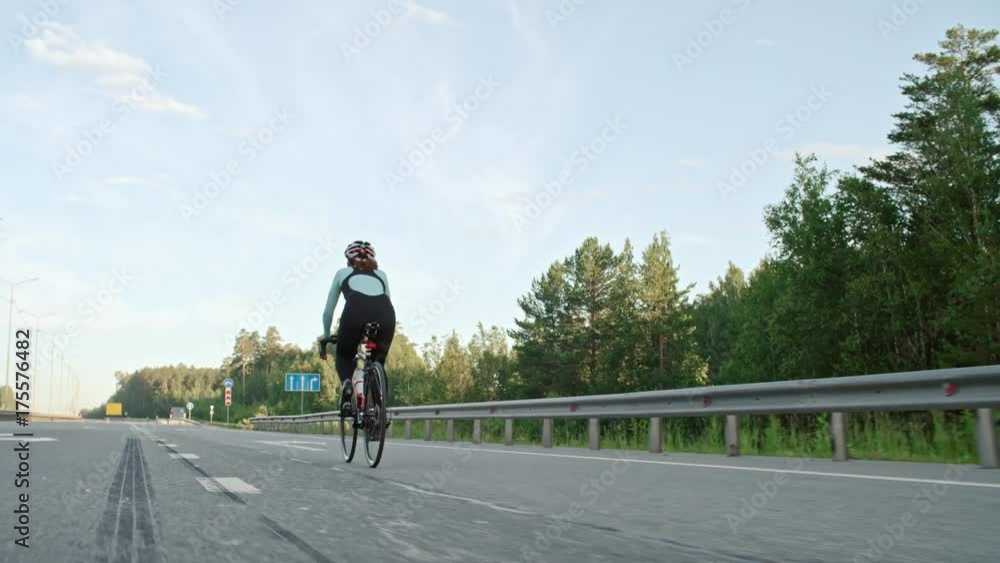 Tracking with rear view of unrecognizable cyclist in professional sportswear and helmet riding bicycle along empty highway Tracking with rear view of unrecognizable cyclist in professional sportswear and helmet riding bicycle along empty highway