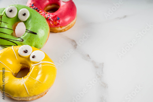 Ideas for children treats on Halloween. Colorful  donuts in the form of monsters with eyes, green, yellow, red chocolate sugar icing. On a white marble table. Copy space © ricka_kinamoto