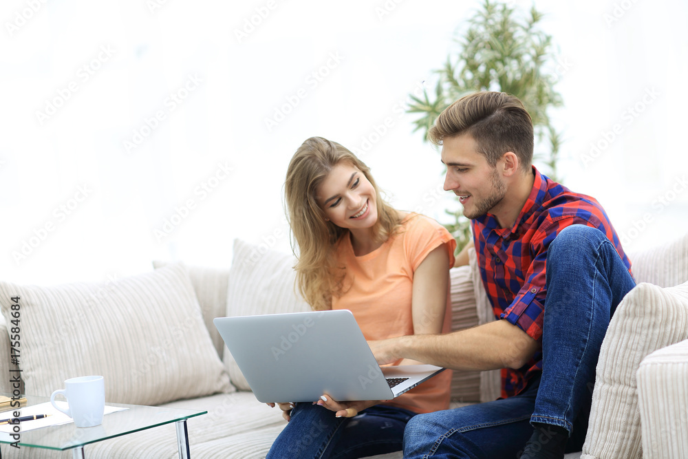 young couple is using a laptop and smiling while sitting on sofa at home