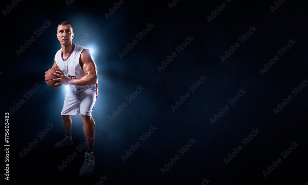 Basketball player stands in the rays of light on a dark background. Player holds a professional basketball ball. Player wears unbranded uniform. Basketball player stands in the rays of light on a dark background. Player holds a professional basketball ball. Player wears unbranded uniform.