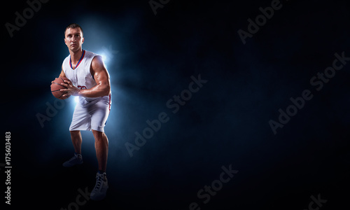 Basketball player stands in the rays of light on a dark background. Player holds a professional basketball ball. Player wears unbranded uniform.