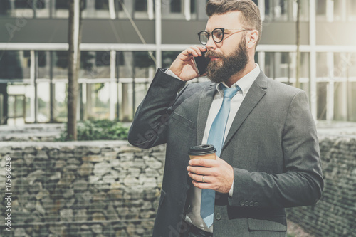 Young businessman in suit and tie is standing outdoor, drinking coffee and talking on his cell phone. In background is modern glass building. Man is working. Negotiations by phone. Business planning.