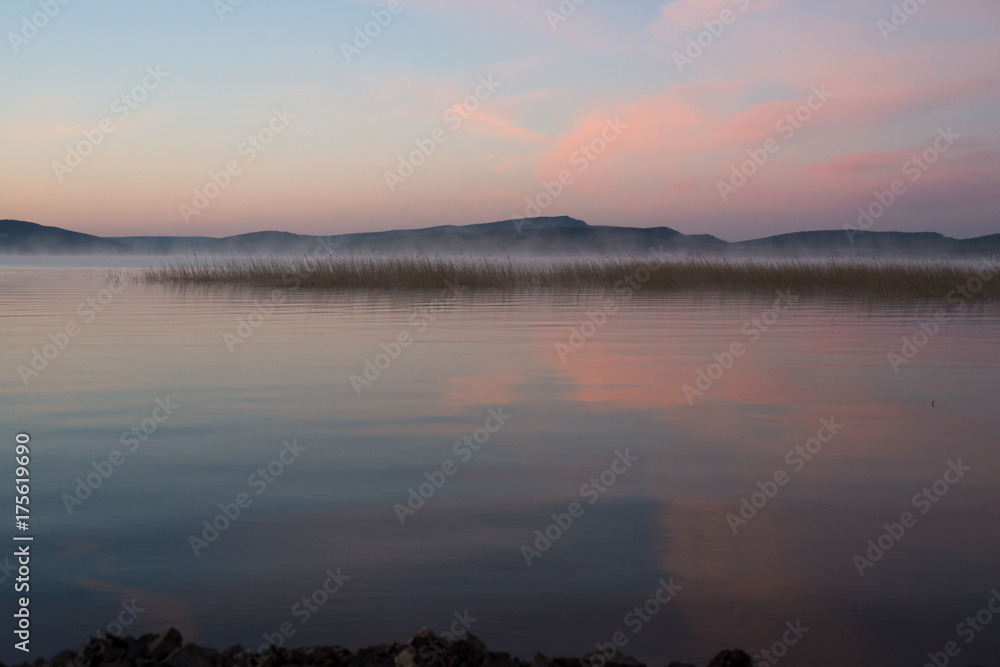 Туманный, ранний, розовый рассвет на озере с горами на заднем плане.
