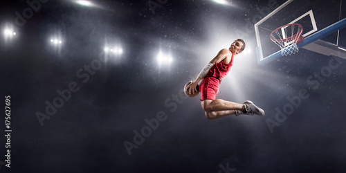 Basketball player makes slam dunk on big professional arena. Player flies through the air with the ball. Player wears unbranded clothes.