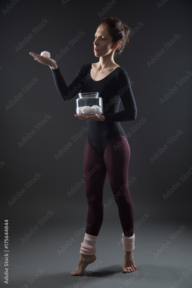 Obraz premium A young slim dancer holds sweet in a dark room after her dance class. Concept idea of a bad habit to be addicted to sweet, sugar. A decision making point - what to choose to make sport or to eat sweet