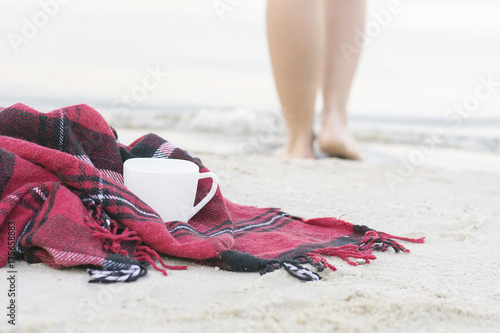 Woman's feet, cup of hot tea and red plaid on the seashore
