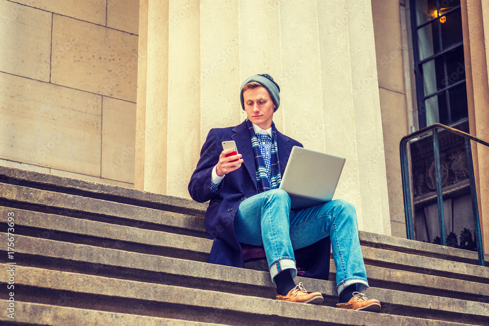 American man traveling, working in New York, wearing blue long woolen overcoat, scarf, jeans, leather shoes, knit hat, sits on stairs outside, works on laptop computer, reads message on cell phone.