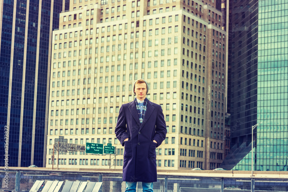 Young American Man traveling in New York, wearing blue long woolen overcoat, scarf, cuffed knit hat, hands in pockets, standing at street park in front of busy business district, looking forward.. Young American Man traveling in New York, wearing blue long woolen overcoat, scarf, cuffed knit hat, hands in pockets, standing at street park in front of busy business district, looking forward..
