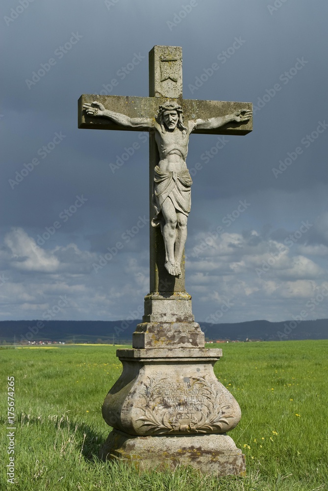 Jesus on the cross, stone statue in a field beneath storm clouds, Hassberge Mountains, Lower Franconia, Bavaria, Germany, Europe