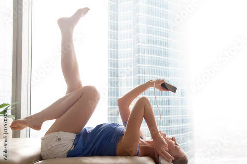 Young woman listening groove music in headphones lying on sofa with feet up, teen girl enjoying new playlist using player app on smartphone, teenager singing along hearing audio track, copy space