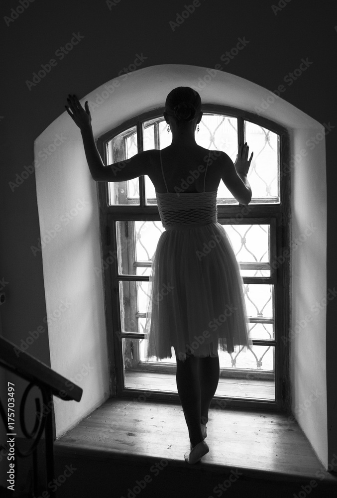 young ballerina in white dress and satin ballet shoes posing on a old window in a dark room. black and white