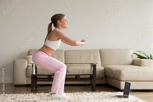 Fotografija Young sporty woman doing two-leg deep squat morning exercise standing in living