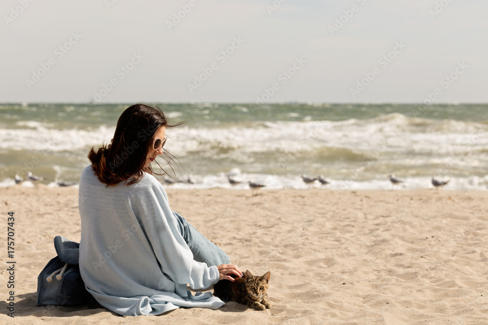 Young woman (brunette) in a light blue cardigan and jeans, with a backpack, sits on the beach and plays with a cat.