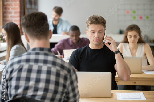 Serious casually dressed businessman talking on cellphone in shared coworking office at work desk in front of laptop. Important deal discussion  business negotiations  client consulting concept.
