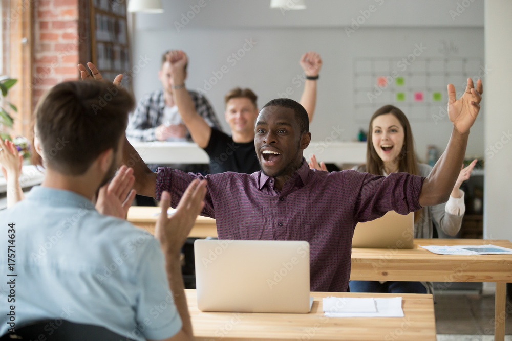 Young african american office worker throws hands in air celebrating achievement at work. Coworkers around cheering and clapping hands. Rewarding outcome, received promotion, achieved success concept.