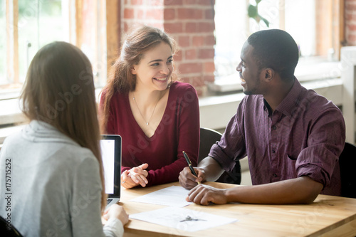 Fotografie Happy multiethnic couple looking at each other before signing contract in front of female real estate agent