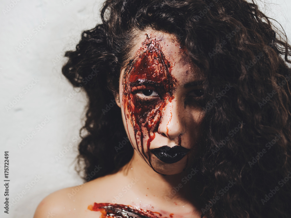 Scary of young zombie girl with Halloween blood makeup. Beautiful latin woman with hair looking into camera in studio. Living dead lady. | Adobe Stock