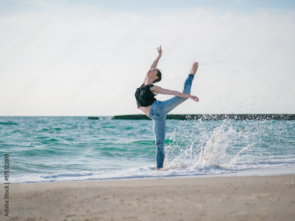 Beautiful scene of a dancing ballerina in black top and jeans on sandy beach ocean or sea in morning. Beautiful brunette woman practicing stretching and exercises.Copy space Beautiful scene of a dancing ballerina in black top and jeans on sandy beach ocean or sea in morning. Beautiful brunette woman practicing stretching and exercises.Copy space
