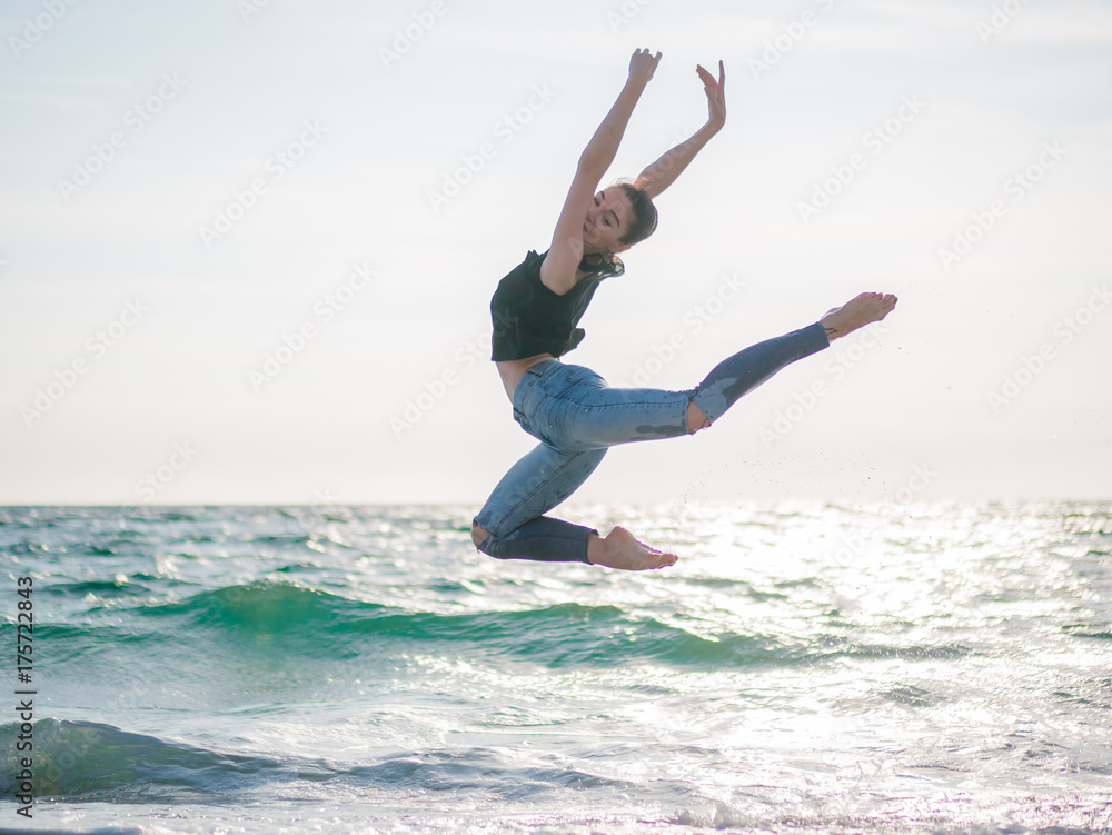 Beautiful scene of a dancing ballerina in black top and jeans on sandy beach ocean or sea in morning. Beautiful brunette woman practicing stretching and exercises.Copy space Beautiful scene of a dancing ballerina in black top and jeans on sandy beach ocean or sea in morning. Beautiful brunette woman practicing stretching and exercises.Copy space