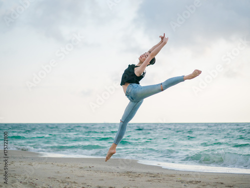 Beautiful scene of a dancing ballerina in black top and jeans on sandy beach ocean or sea in morning. Beautiful brunette woman practicing stretching and exercises.Copy space