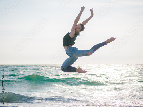 Beautiful scene of a dancing ballerina in black top and jeans on sandy beach ocean or sea in morning. Beautiful brunette woman practicing stretching and exercises.Copy space