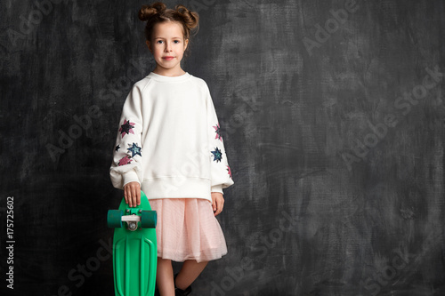 Fashionable little girl in a blank white sweatshirt with a skate stands in full growth against a chalkboard background