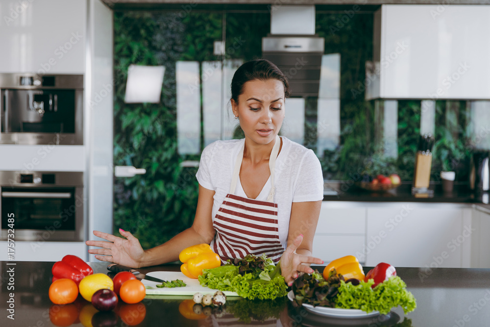 A young confused and thoughtful woman in apron decides what to cook in the kitchen. Healthy food - Vegetable salad. Diet. Dieting concept. Healthy lifestyle. Cooking at home. Prepare food