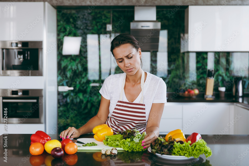 A young confused and thoughtful woman in apron decides what to cook in the kitchen. Healthy food - Vegetable salad. Diet. Dieting concept. Healthy lifestyle. Cooking at home. Prepare food