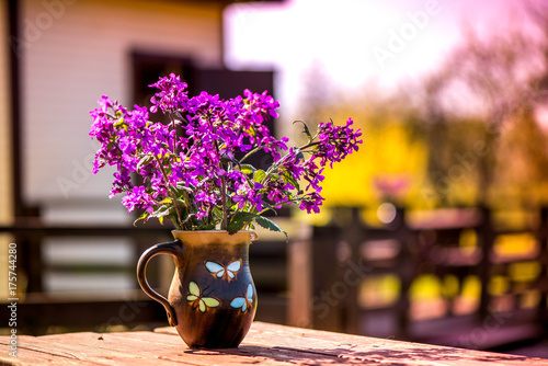 Early summer time. Macro shot of lilac flowers in vase standing on wooden, rustic, vintage table in middle of garden on early morning. Sun is up, perfect day for outdoor activities. Breakfast outside