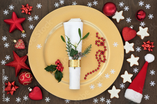 Christmas table place setting with gold dinner plate, napkin with cedar, mistletoe and holly, santa hat, gingerbread biscuits and bauble decorations on oak wood background.