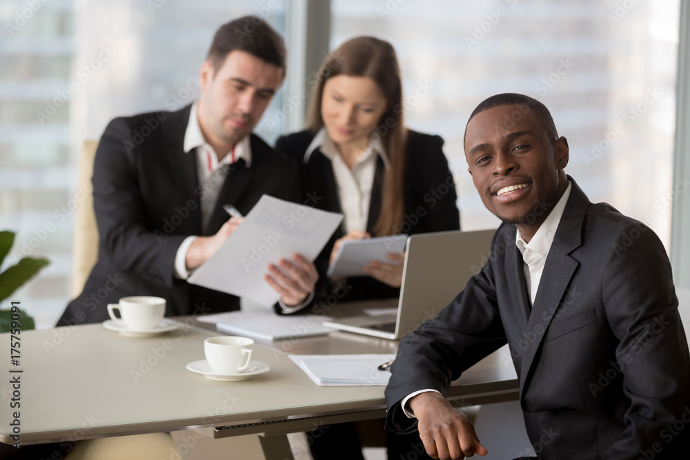 Portrait of confident african american millennial businessman sitting at desk, caucasian colleagues working on document at background. Multinational financial analysts examining company annual report Portrait of confident african american millennial businessman sitting at desk, caucasian colleagues working on document at background. Multinational financial analysts examining company annual report