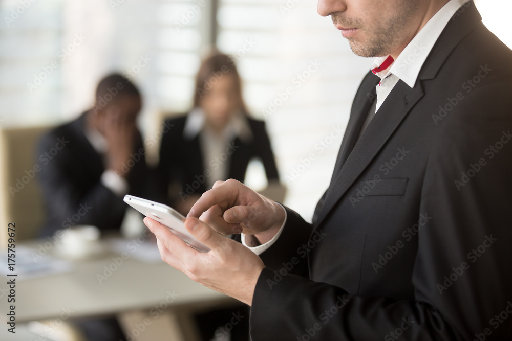 Φωτογραφία Close up image of businessman tapping on cellphone screen with finger, dialing number, writing message, man and woman in formal wear sitting at desk in background