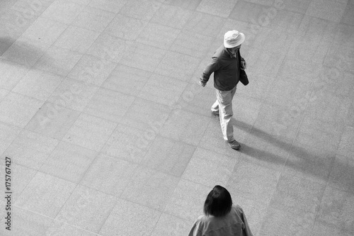 Man in a white hat and jacket and an Asian woman walk through a train station in Europe.  Looking down on two people from above, black and white image.