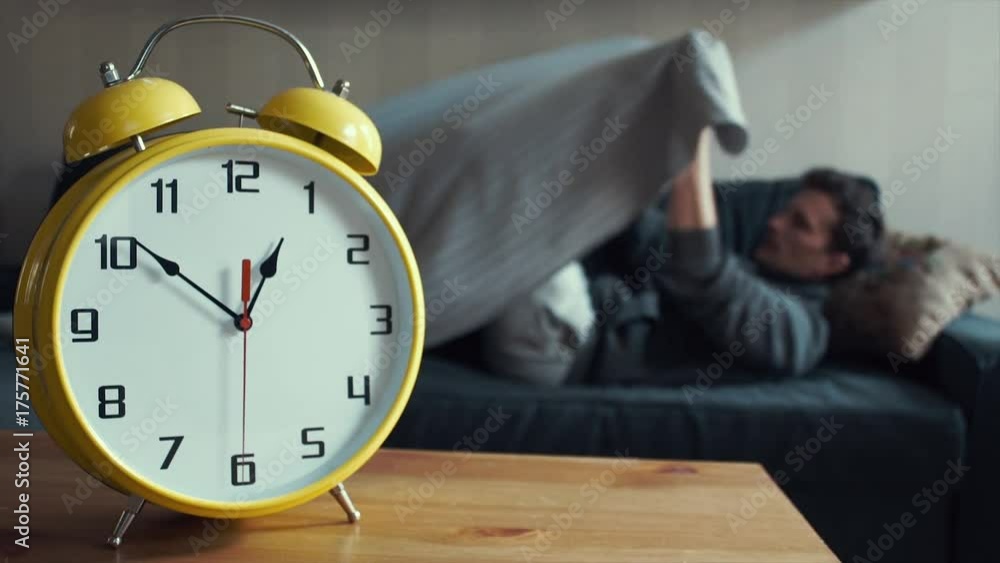 Young man lies covered with blanket on couch at home. Big yellow clock ticking Young man lies covered with blanket on couch at home. Big yellow clock ticking