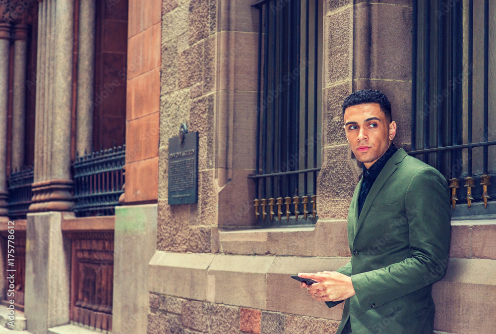 American Businessman traveling in New York, wearing green suit, standing against vintage wall with windows on street, taking work break, texting on cell phone, looking around, thinking.